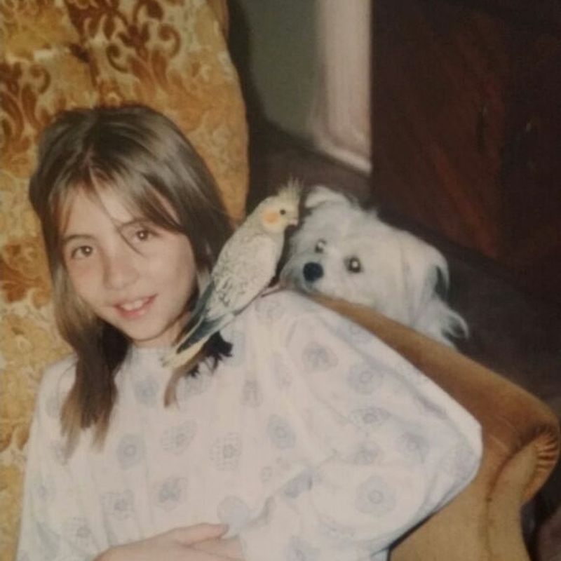 A young Camila with a cockatiel perched on her shoulder and a small white dog beside her.